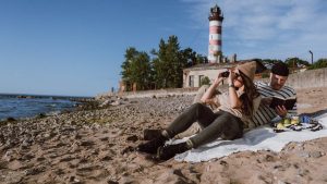 A couple enjoys a beach day by a lighthouse, with binoculars and a book for recreation.