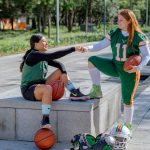 Two female athletes share a fist bump while sitting outdoors, holding sports equipment in Mexico City.